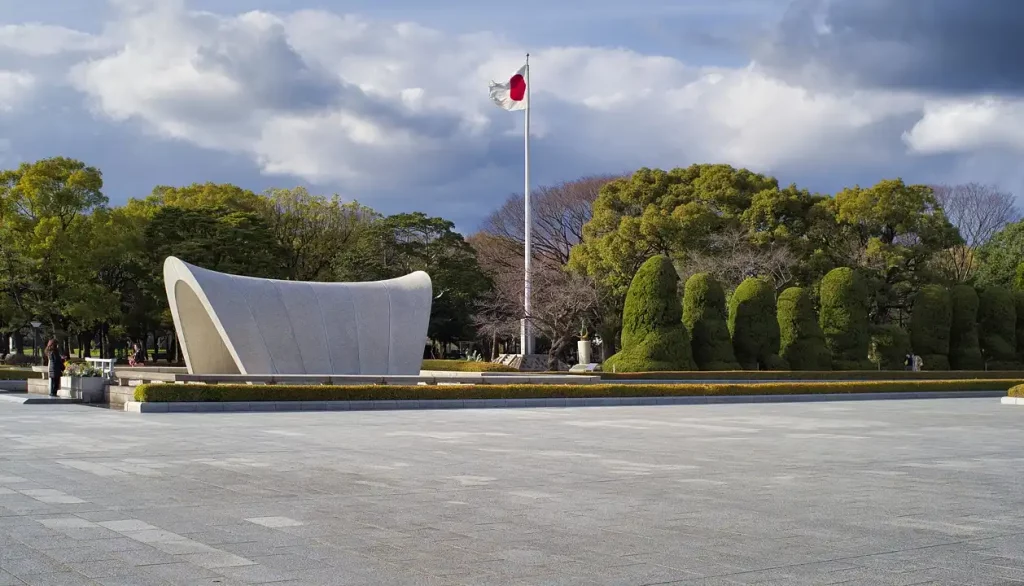 Monumento no Parque da Paz em Hiroshima com a bandeira japonesa ao fundo em um dia parcialmente nublado.