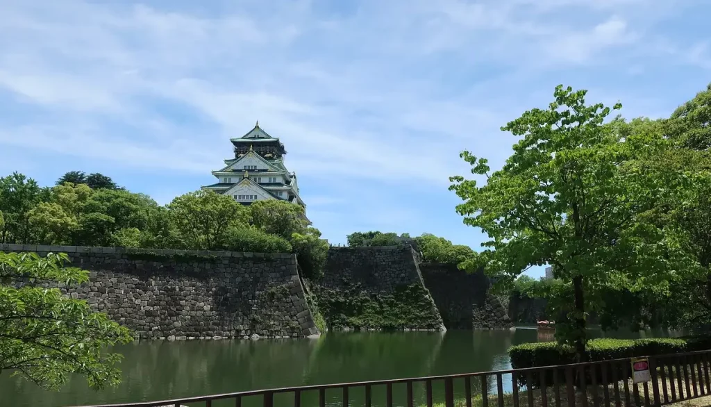 Vista do Castelo de Osaka cercado por árvores verdes e refletido no fosso, em um dia ensolarado.