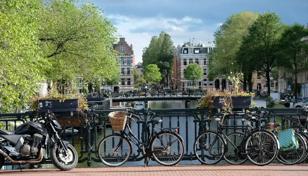 Bicicletas e uma motocicleta estacionadas em uma ponte sobre um canal em Amsterdã, com árvores e prédios ao fundo, mostrando a popularidade do uso de bicicletas como meio de transporte na Holanda.