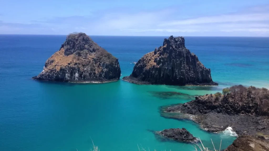 Imagem de duas formações rochosas icônicas emergindo do mar azul-turquesa em Fernando de Noronha, Brasil, com um céu limpo e uma costa rochosa.