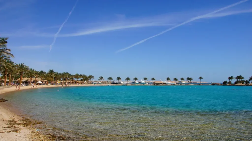 Uma praia tranquila no Mar Vermelho, Egito, com águas azuis transparentes, palmeiras ao longo da costa, e barcos de lazer ancorados ao fundo sob um céu claro e ensolarado.