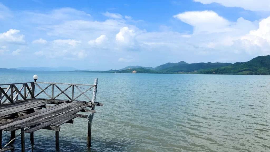 Um cenário sereno do Mar de Andaman, na Tailândia, com um cais de madeira simples que se estende sobre a água calma, cercado por montanhas ao fundo e sob um céu azul claro.