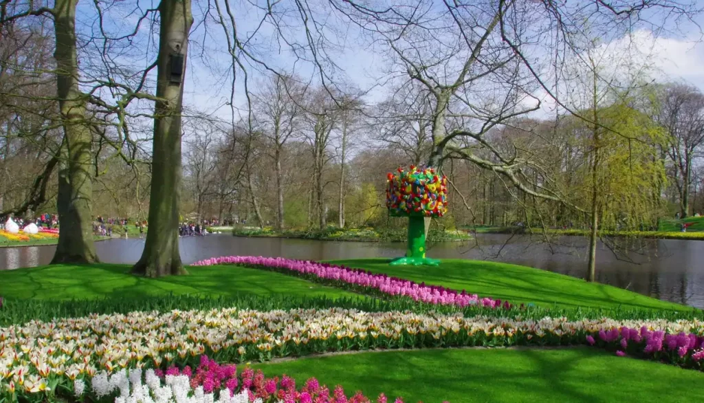 Uma vista panorâmica de um belo jardim com tulipas coloridas em plena floração, com um lago e árvores ao fundo, representando a beleza dos parques na Holanda durante a primavera.