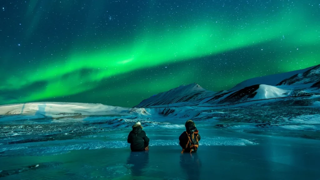 Dois viajantes sentados em um lago congelado, observando a aurora boreal brilhante iluminando o céu estrelado, com montanhas nevadas ao fundo.