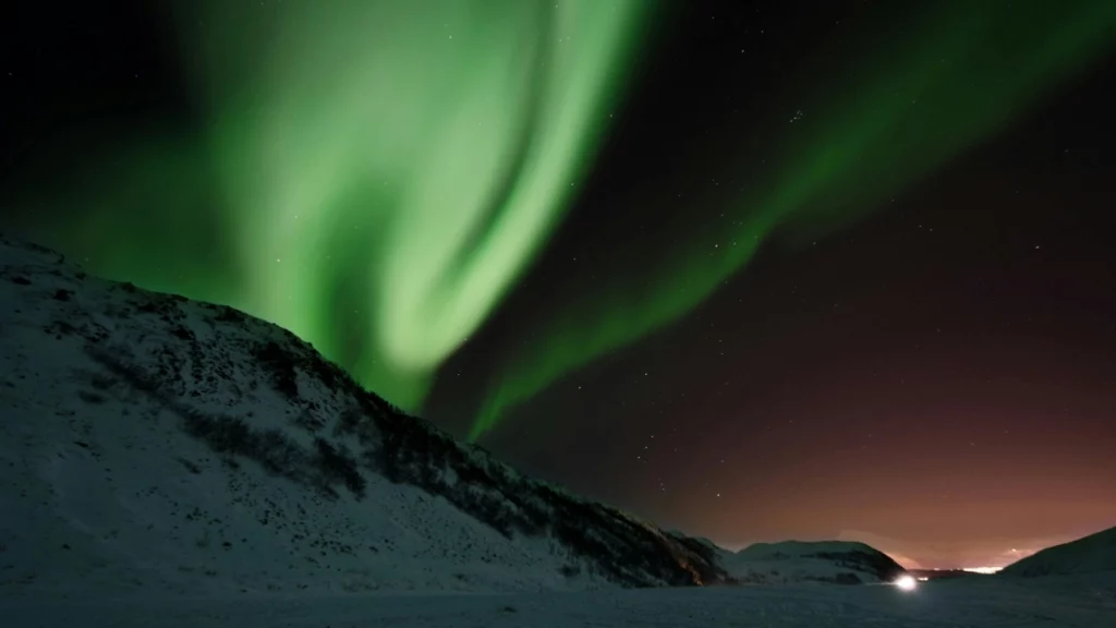 Aurora boreal verde e vibrante dançando sobre uma paisagem montanhosa coberta de neve, com um brilho leve no horizonte, criando um contraste de cores no céu escuro.