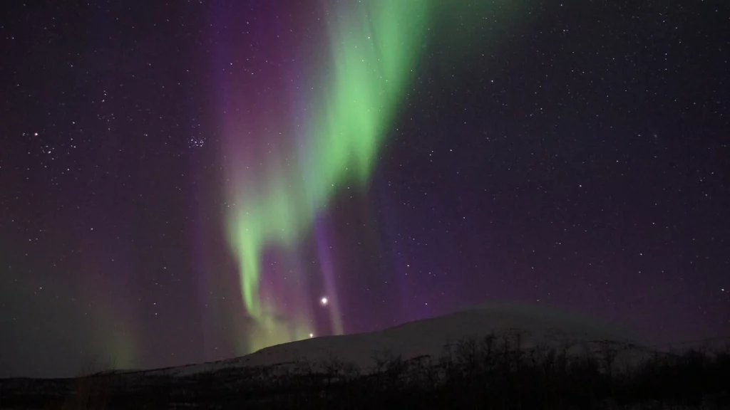 Aurora boreal nas cores verde e roxa iluminando o céu noturno acima de uma montanha, proporcionando um espetáculo natural impressionante.