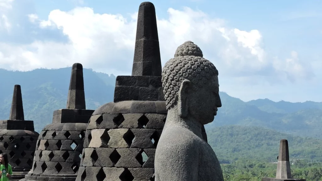 Imponente estátua de Buda em Borobudur, o maior templo budista do mundo, rodeada por estatuetas em pedra com uma paisagem montanhosa ao fundo.