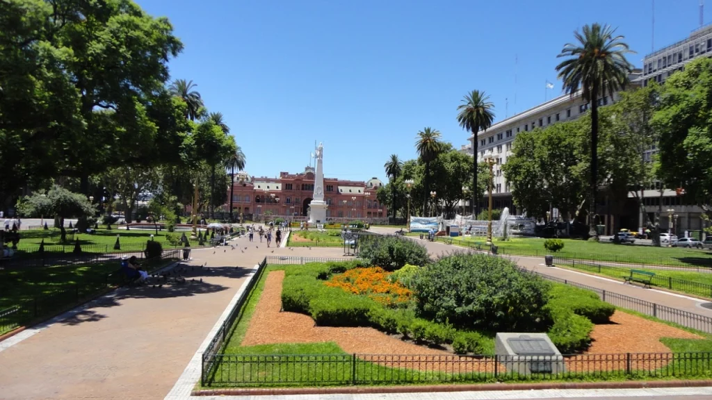 Praça de Maio em Buenos Aires com a Casa Rosada ao fundo, cercada por jardins floridos e palmeiras sob um céu claro.