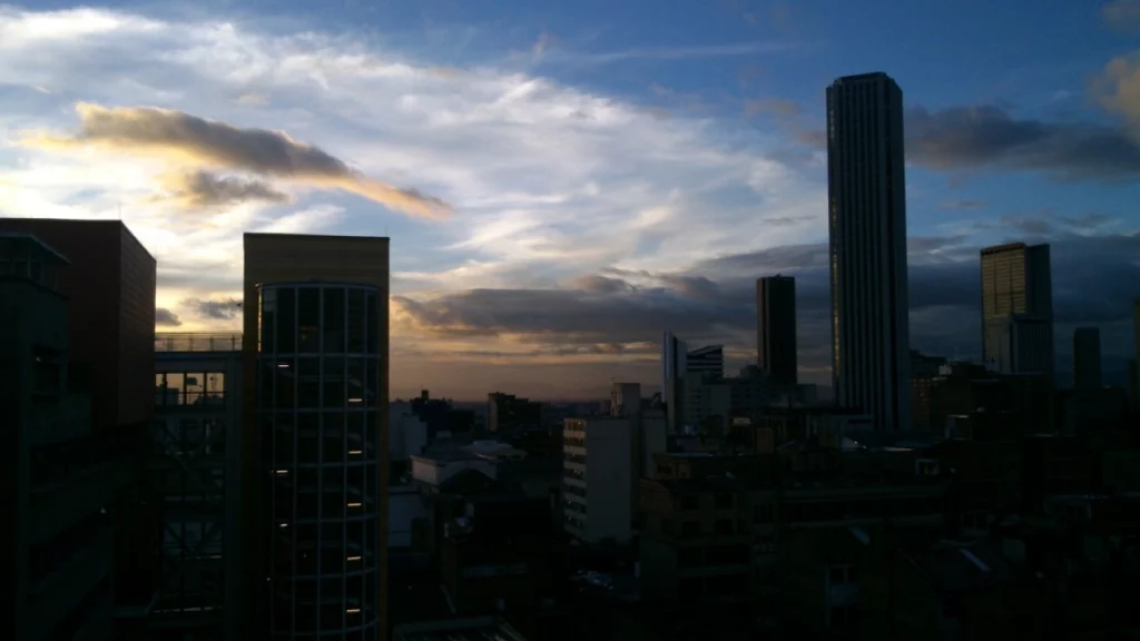 Vista de uma cidade ao entardecer, com arranha-céus altos contrastando contra o céu nublado e colorido pelo pôr do sol.