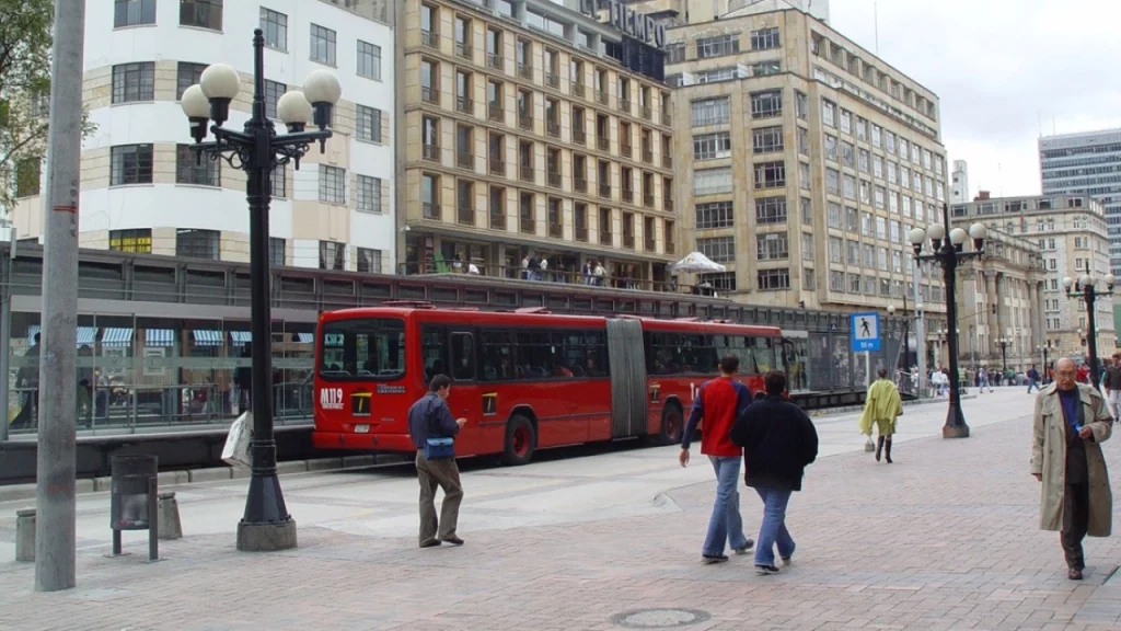 Cena urbana movimentada com pessoas caminhando em uma rua larga, ônibus vermelho em uma estação de parada e edifícios antigos ao fundo.