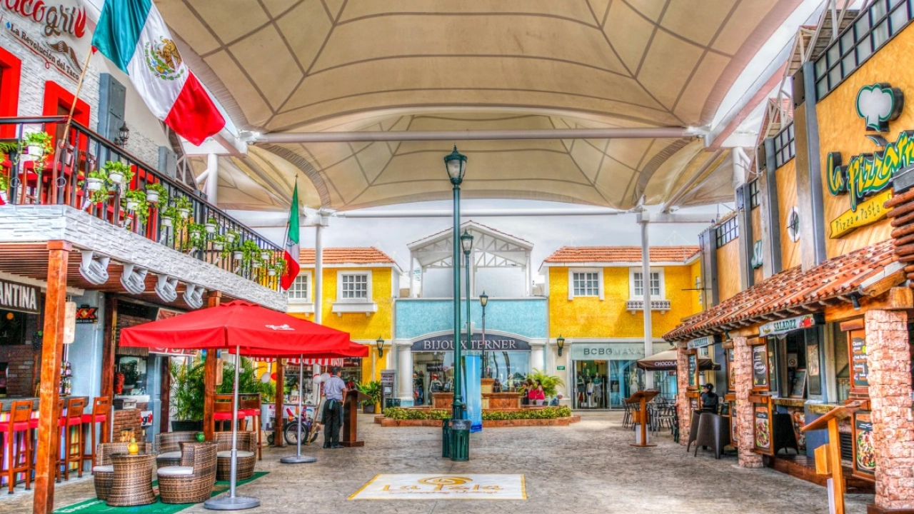 Plaza al aire libre con restaurantes en Cancún, México, adornada con banderas mexicanas y rodeada de tiendas locales, ideal para compras y comida típica.