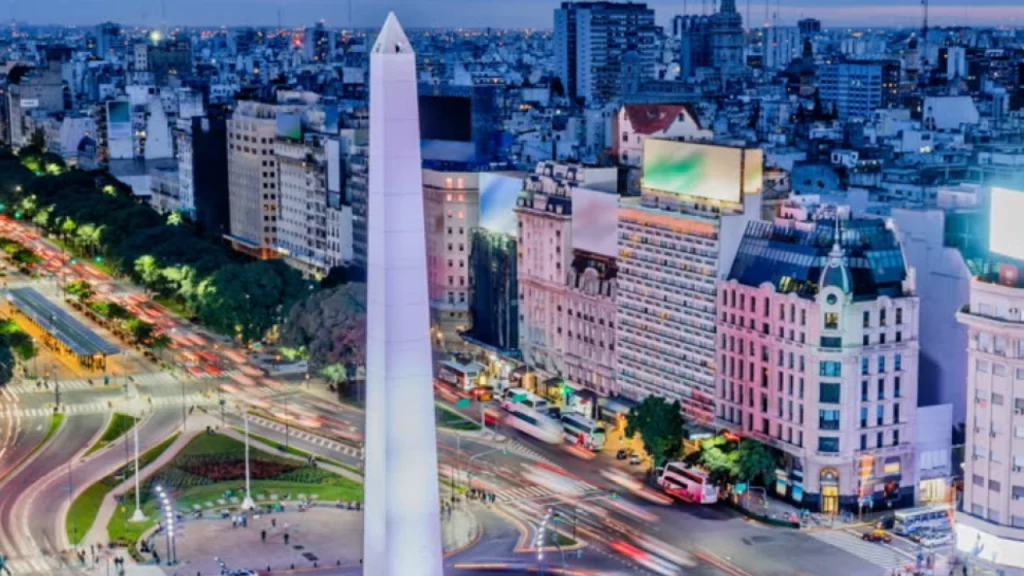 Vista noturna do Obelisco de Buenos Aires, rodeado por luzes de carros em movimento e edifícios iluminados no centro da cidade.