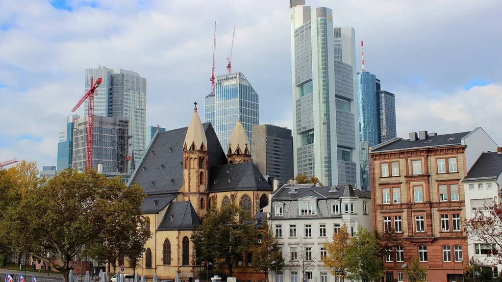 contraste entre antigo e moderno): "Uma visão de Frankfurt mostrando o contraste entre a arquitetura histórica de igrejas e os modernos arranha-céus do distrito financeiro.