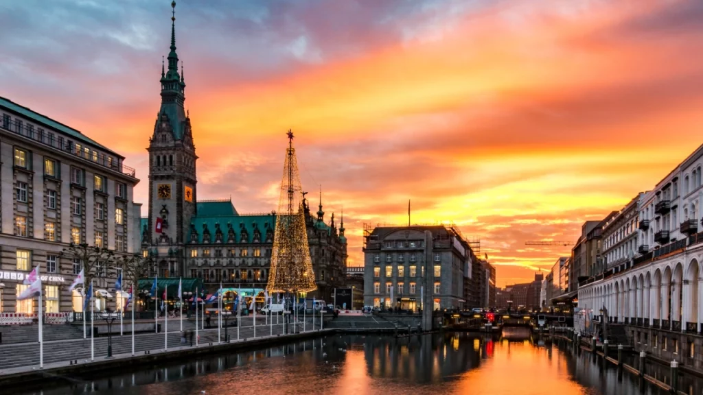 Vista deslumbrante do entardecer em Hamburgo, Alemanha, com as luzes da cidade refletindo no canal e uma árvore de Natal iluminada em frente ao imponente prédio da prefeitura.