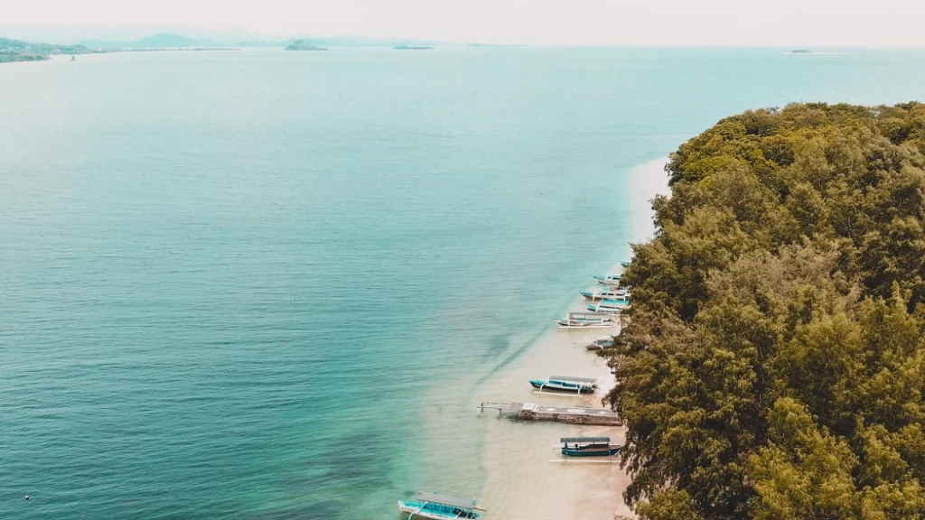 Barcos coloridos estacionados ao longo de uma praia de areia branca na costa da Indonésia, com águas calmas e cristalinas que refletem o céu claro.