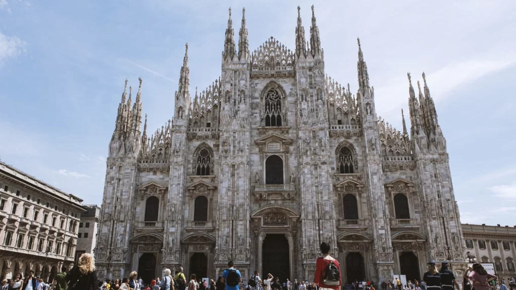 Fachada imponente da Catedral de Milão, com turistas caminhando pela praça. Um dos maiores exemplos de arquitetura gótica na Europa.