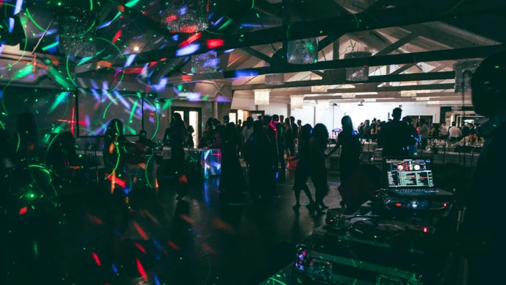 Um ambiente de festa, com luzes de discoteca coloridas e pessoas dançando em um salão enquanto um DJ toca música ao fundo.