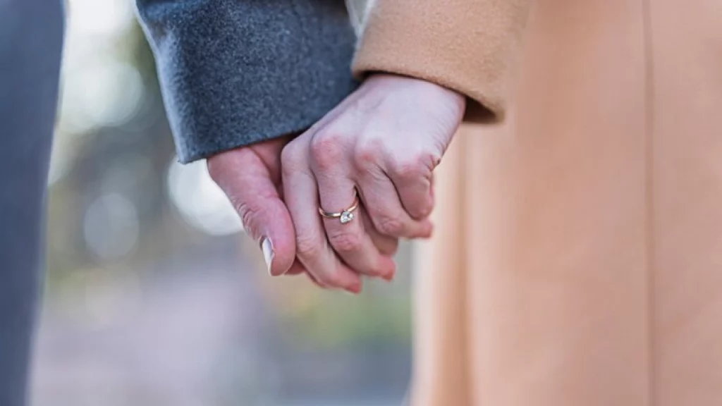 Um close-up de duas pessoas de mãos dadas, uma delas com um anel de casamento visível, simbolizando afeto e compromisso.