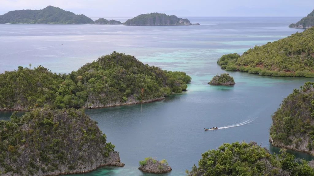 Vista aérea de pequenas ilhas verdes cercadas por águas cristalinas em Raja Ampat, Indonésia, com um barco navegando tranquilamente entre as ilhas.