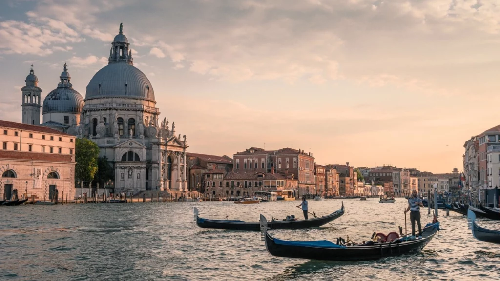 Vista do Grande Canal em Veneza ao pôr do sol, com gôndolas navegando em frente à Basílica di Santa Maria della Salute, capturando a atmosfera romântica e histórica da cidade.