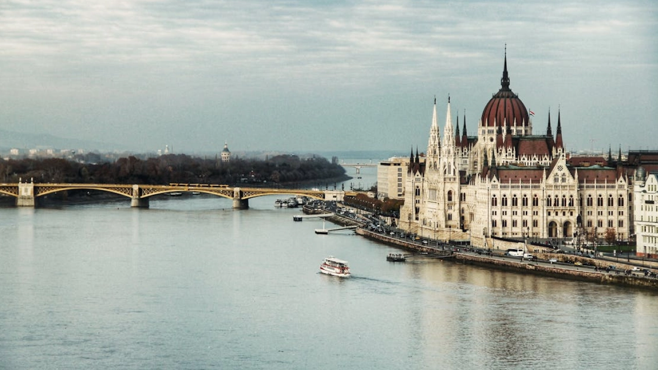 Vista do edifício do Parlamento de Budapeste ao lado do Rio Danúbio, com uma ponte ao fundo e um barco navegando no rio.