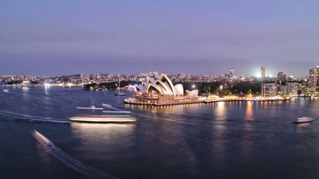  A icônica Sydney Opera House iluminada à noite, com barcos cruzando a baía e a cidade vibrante ao fundo.