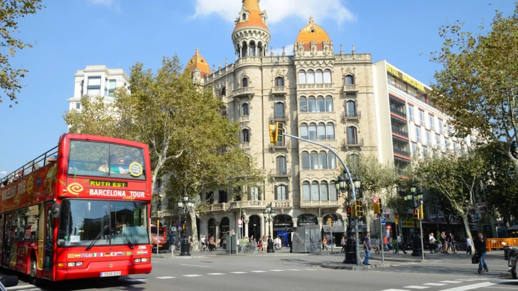 Ônibus turístico de dois andares passando por uma avenida movimentada em Barcelona, com edifícios históricos ao fundo.