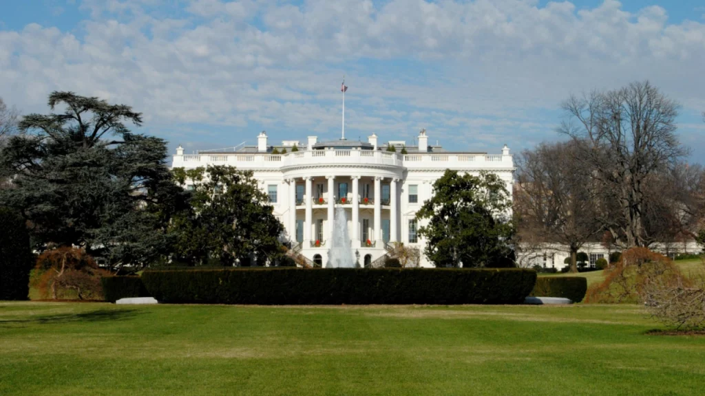 Imagem frontal da Casa Branca em Washington, D.C., cercada por árvores e grama verde, com o céu parcialmente nublado ao fundo.