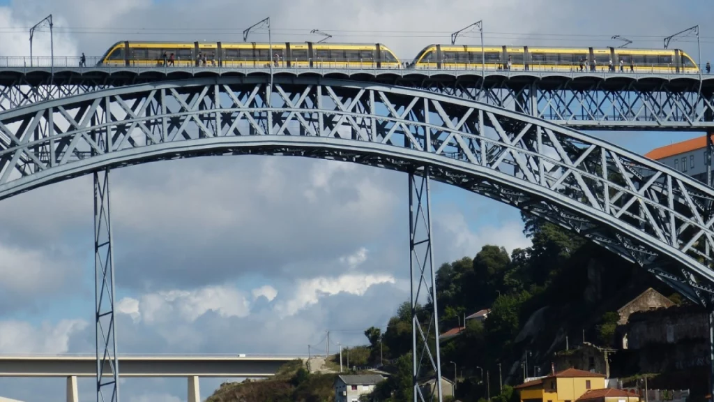 Trem amarelo atravessando a Ponte Luís I, um ícone da cidade do Porto, em Portugal, com o céu azul e nuvens ao fundo.