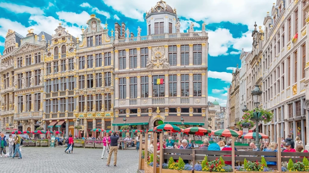 Fachada ornamentada de edifícios históricos na Grand Place de Bruxelas, cercada por mesas ao ar livre e turistas apreciando o ambiente.