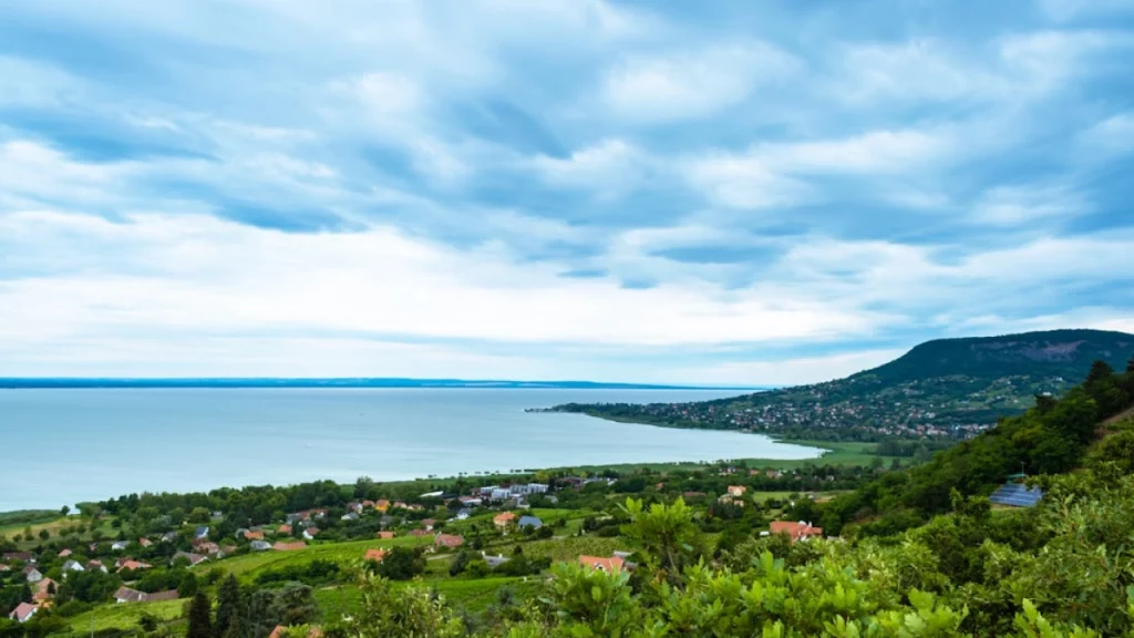 Vista panorâmica de uma vila à beira de um lago, cercada por colinas verdes e céu nublado.