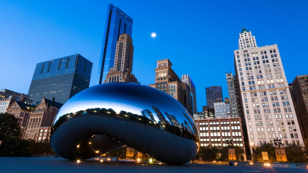 O famoso "feijão" espelhado (Cloud Gate) de Chicago reflete os edifícios iluminados da cidade sob um céu azul durante o entardecer.