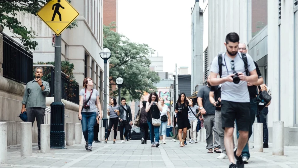 Várias pessoas caminhando em uma rua movimentada com prédios ao redor, algumas tirando fotos e outras mexendo no celular.