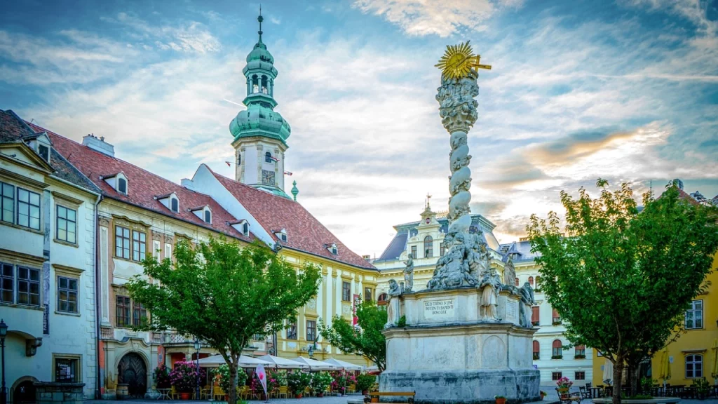 Praça com uma coluna decorativa no centro, rodeada por árvores e prédios históricos coloridos com uma torre ao fundo.