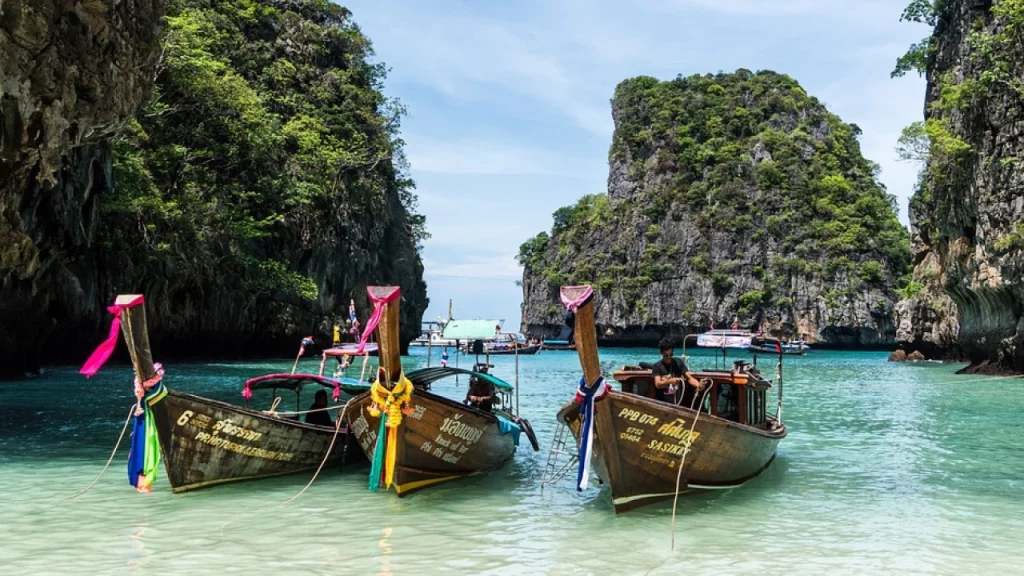 Barcos tradicionais tailandeses ancorados em uma praia de águas cristalinas cercada por penhascos verdes na Tailândia.