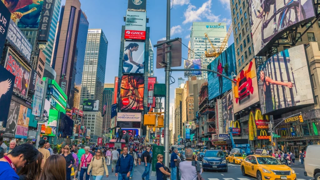 Foto de Times Square em Nova York, com muitas pessoas andando pelas calçadas e cruzando a rua, cercadas por prédios altos e grandes painéis publicitários iluminados.