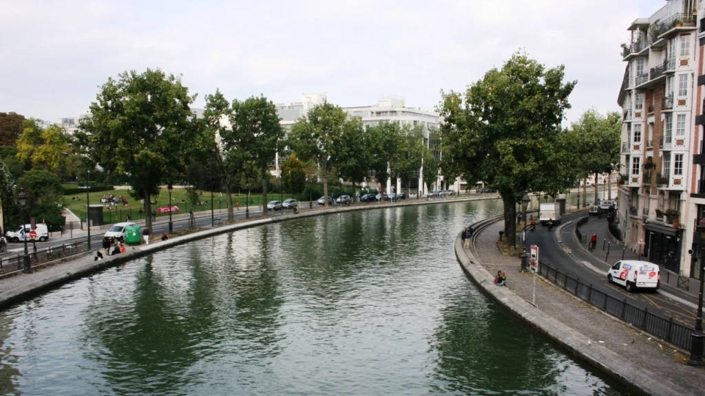 Canal sinuoso cercado por árvores e edifícios em Paris, com pessoas sentadas na margem, aproveitando o ambiente tranquilo e urbano.