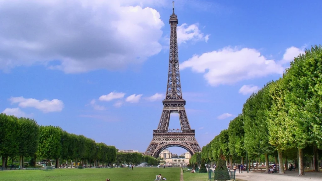 Torre Eiffel vista dos jardins de Champ de Mars, com árvores alinhadas ao longo do caminho e pessoas relaxando na grama em um dia ensolarado.