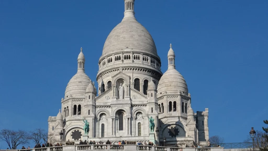 A Basílica de Sacré-Cœur, em Montmartre, Paris, com suas cúpulas brancas impressionantes contra um céu azul claro, e turistas reunidos na escadaria.