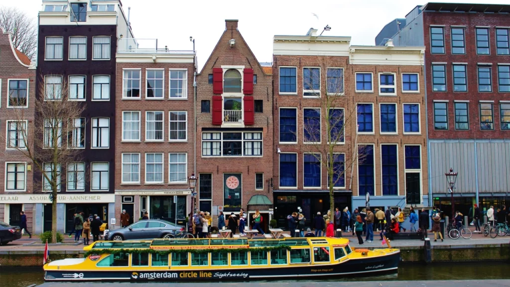 Fachada da Casa de Anne Frank em Amsterdã, com um barco turístico atracado no canal em frente.