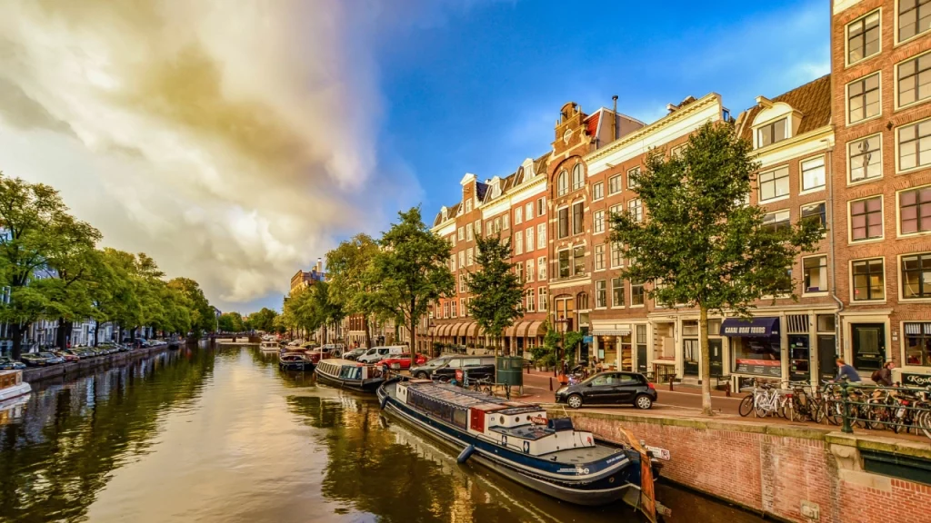 Vista dos tradicionais canais de Amsterdã ao pôr do sol, com casas típicas e barcos-casa atracados, sob um céu parcialmente nublado.