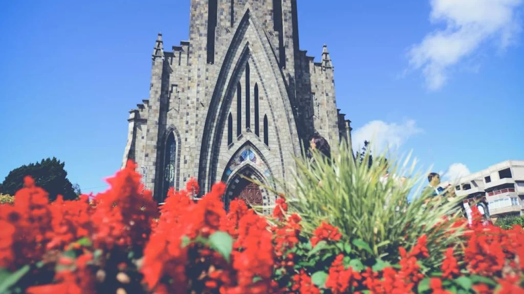 Fachada da Catedral de Pedra em Canela, RS, destacada por flores vermelhas no primeiro plano e um céu azul ao fundo.