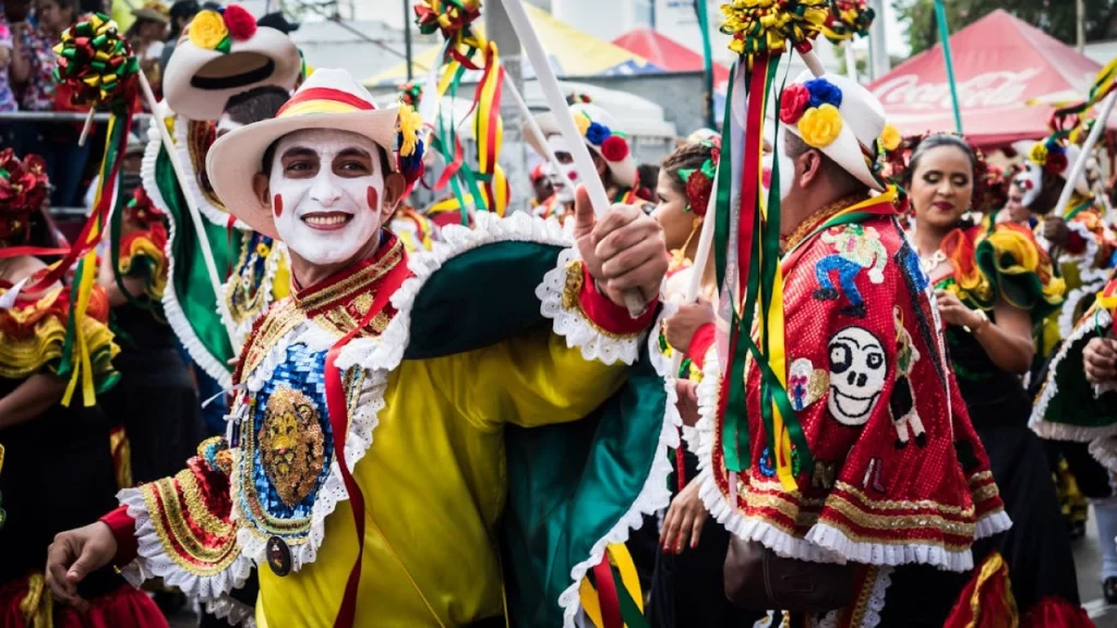 Desfile do Carnaval de Barranquilla, Colômbia, com foliões vestindo roupas coloridas e máscaras vibrantes. Um homem com o rosto pintado sorri e segura um bastão branco, enquanto outros participantes usam chapéus decorados e trajes tradicionais."