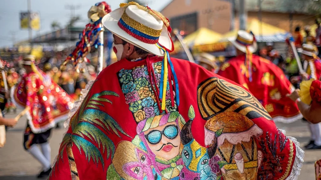 Participante do Carnaval de Barranquilla vestindo um manto vermelho bordado com desenhos coloridos e brilhantes, incluindo figuras folclóricas e símbolos da cultura colombiana. Ele segura um bastão enquanto desfila pelas ruas lotadas de espectadores.