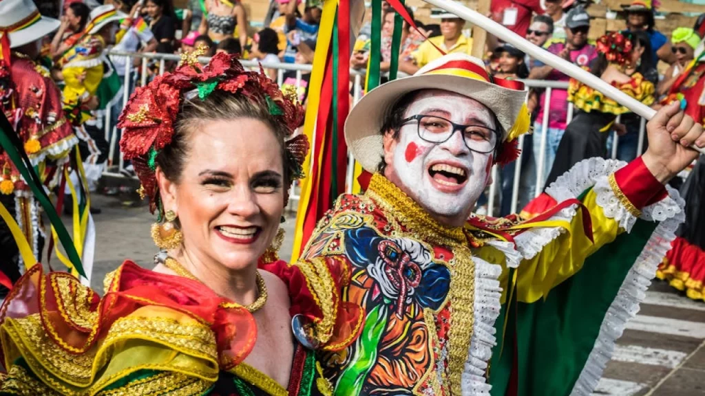 Casal sorridente no Carnaval de Barranquilla, vestindo trajes tradicionais coloridos e decorados. O homem tem o rosto pintado de branco e usa um chapéu enfeitado, enquanto a mulher exibe um vestido vibrante com detalhes dourados e flores nos cabelos.