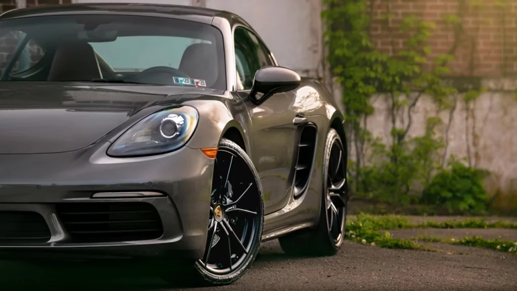 Carro esportivo Porsche prateado estacionado ao lado de um prédio de tijolos antigos, com vegetação crescendo ao fundo.