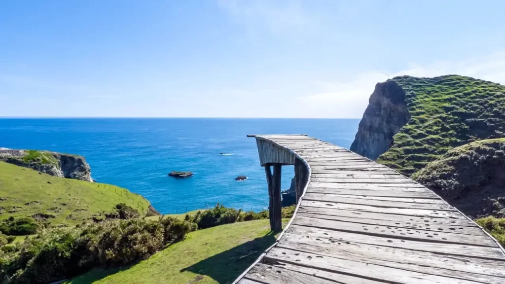 Vista da Isla de Chiloé, no Chile, com passarela de madeira sobre as falésias, mar azul intenso e colinas verdes.