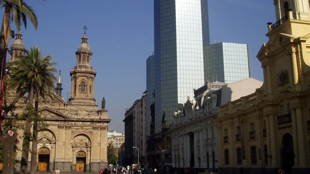 Fachada da Catedral Metropolitana de Santiago vista da Plaza de Armas, com palmeiras e prédios históricos ao redor no centro da capital chilena.