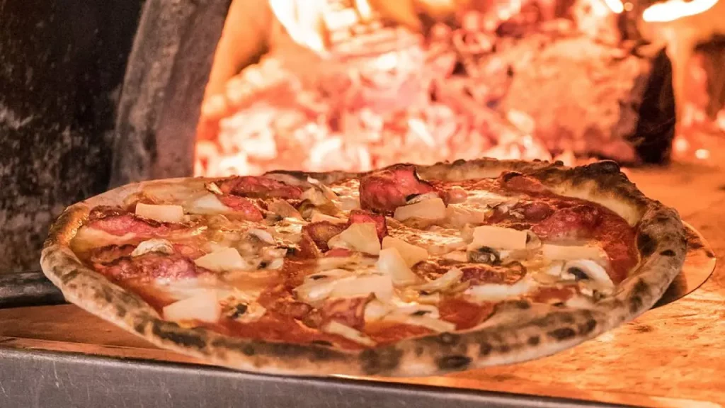 Pizza de salame, cogumelos e queijo dentro de forno a lenha com brasas acesas.