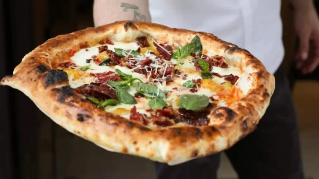 Pizza artesanal de borda alta com tomate seco, pimentão amarelo, queijo e folhas de manjericão, sustentada por braço tatuado de pizzaiolo.